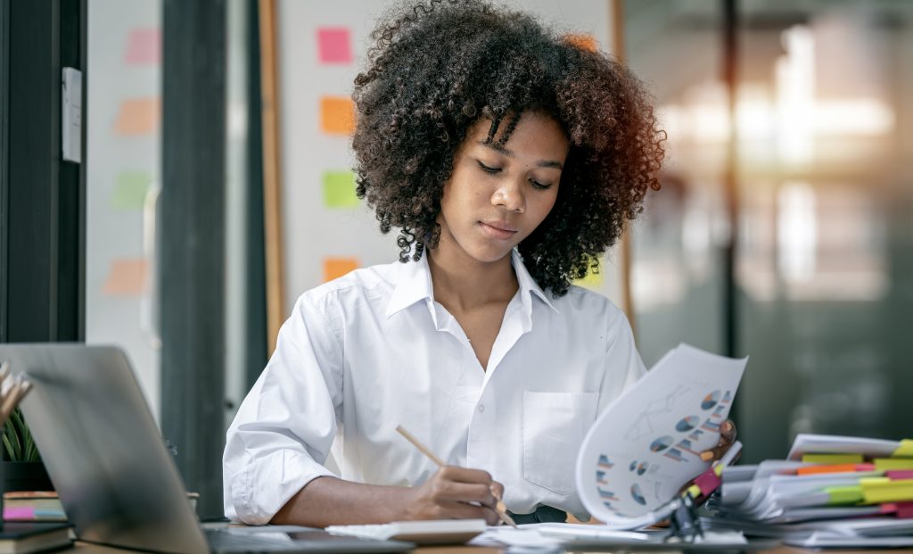 Attractive young african american business woman working on business graph chart, analysing research finance and marketing at office desk.