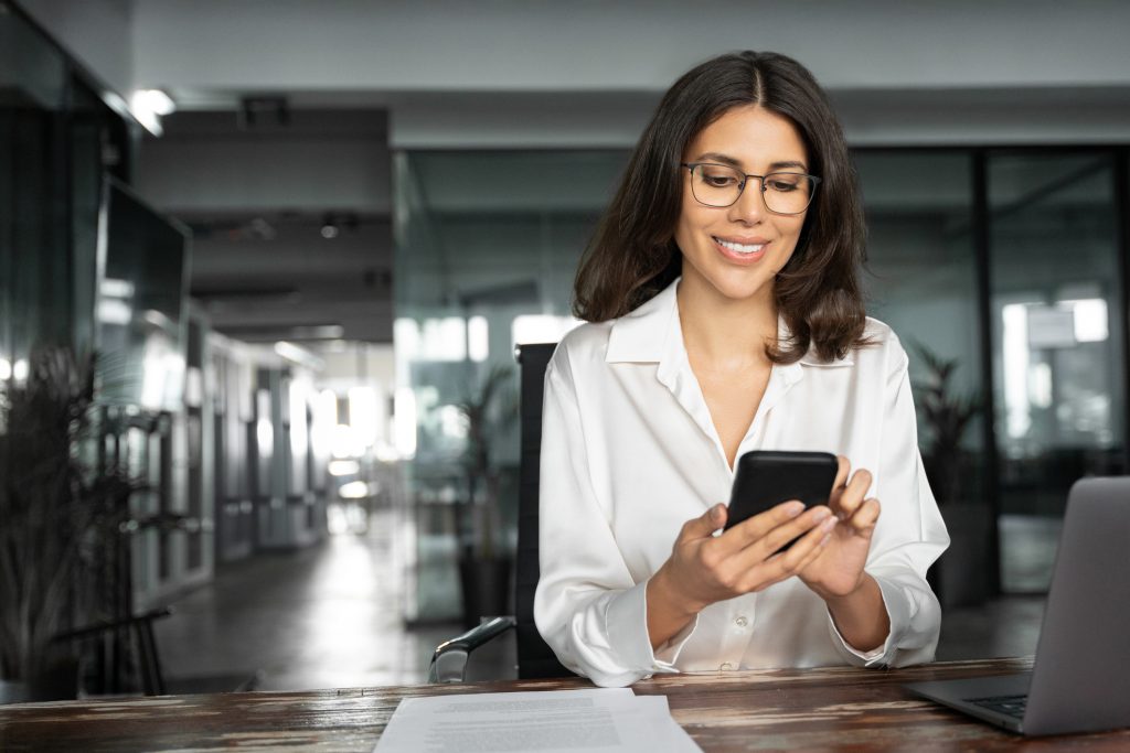 Middle aged Hispanic business manager ceo using cell phone mobile app, laptop. Smiling Latin or Indian mature woman businesswoman holding smartphone sit in office working online on gadget, copy space.